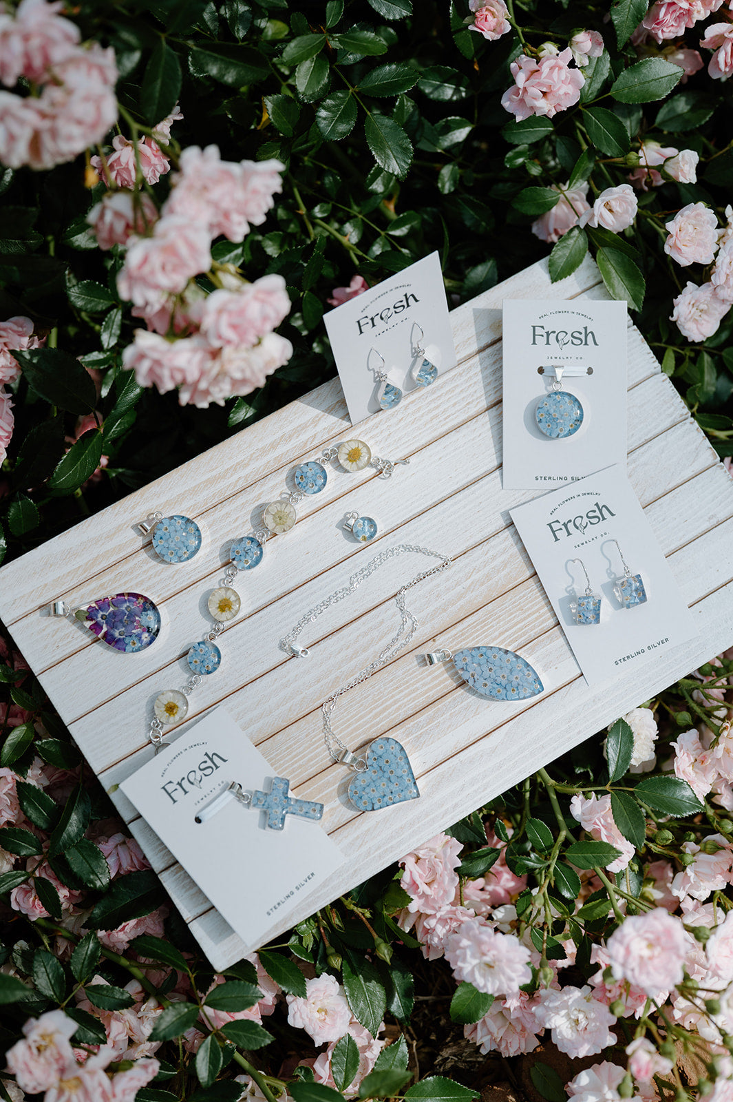 Jewelry set with 'Fresh' branding on a wooden display board, surrounded by pink flowers.