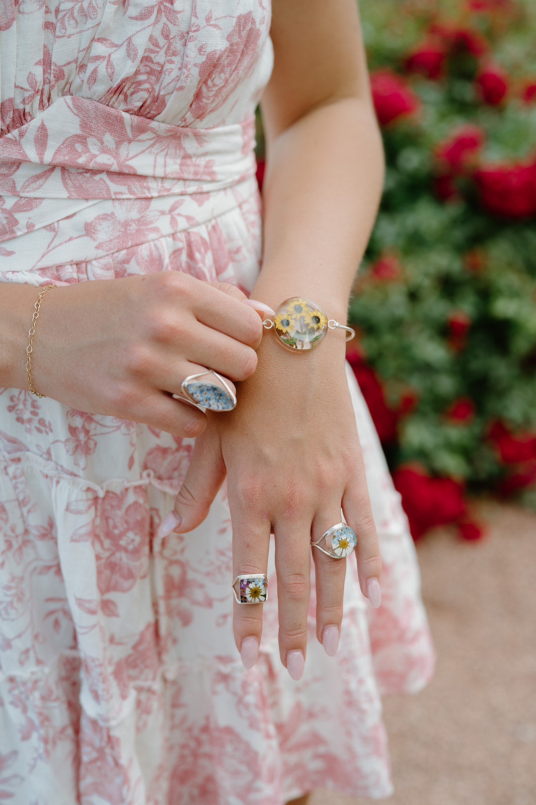 Close-up of a person wearing multiple rings with a floral dress and blurred red flowers in the background