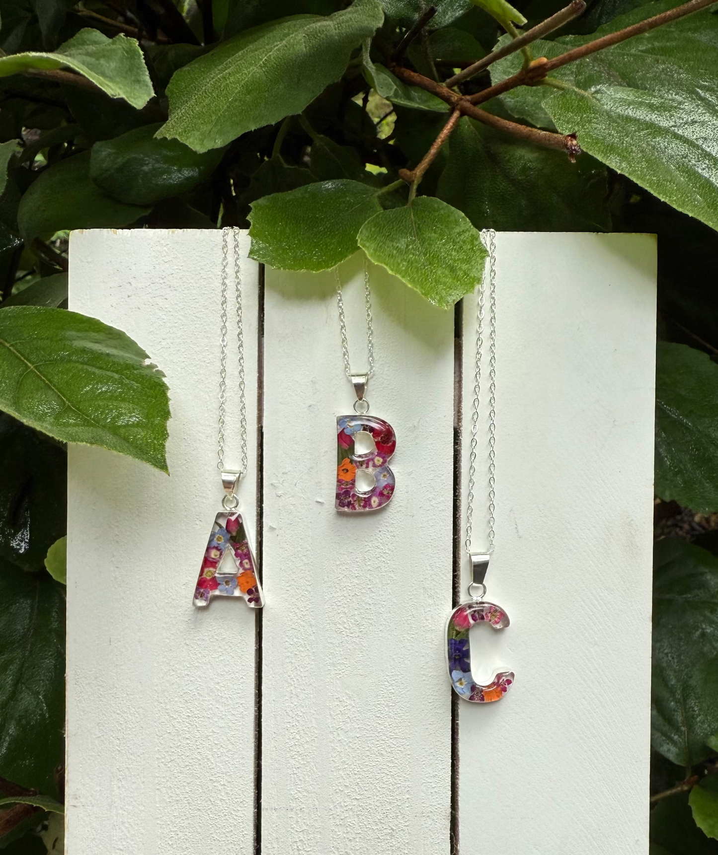Three colorful letter pendants on silver chains against a white wooden background with green leaves.