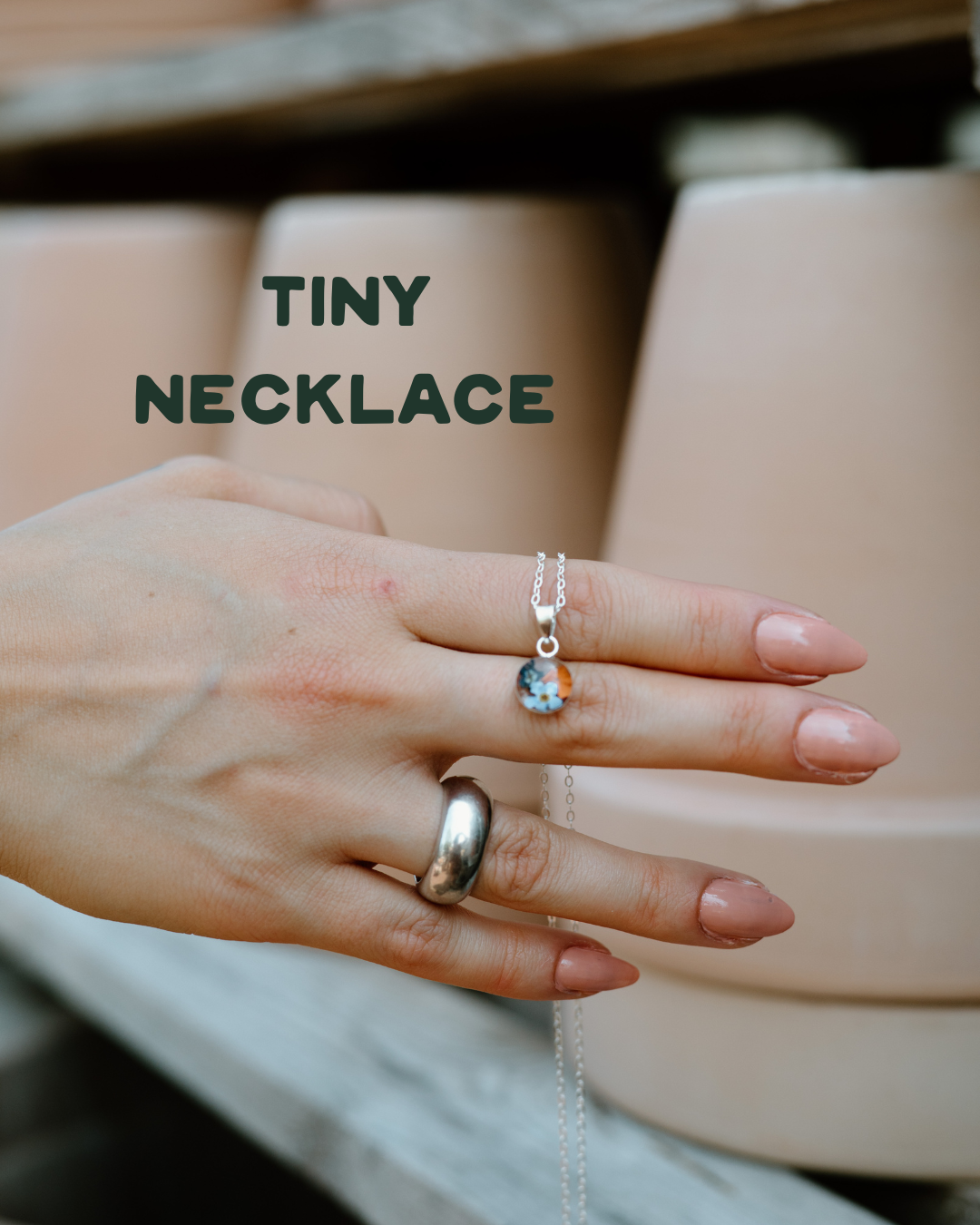 Hand wearing a silver ring and holding a tiny necklace with wildflowers, set against a neutral background.