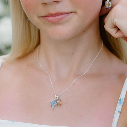 Young girl with blonde hair wearing a white dress with blue floral patterns, standing outdoors.