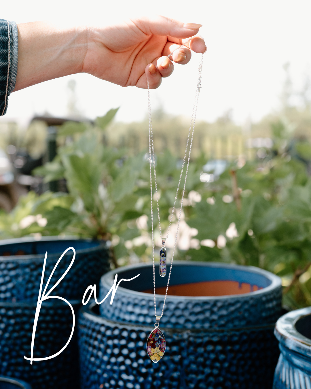 Hand holding a necklace with a pendant in front of potted plants
